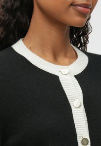 Cardigan noir avec un col et une patte de boutonnage côtelés blancs, ornés de boutons ronds dorés. Texture douce avec un gros plan sur le décolleté.