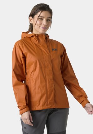 Mujer con chaqueta impermeable con capucha naranja y pantalones de exterior gris oscuro, de pie contra un fondo liso.