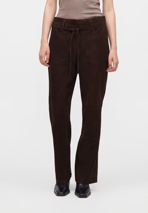 Personne portant un pantalon taille haute marron foncé avec une ceinture en tissu, un haut beige sans manches et des chaussures noires à bout carré, debout devant un fond uni.