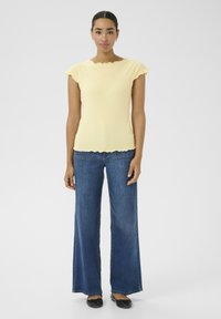 Un haut jaune à côtes avec un col festonné et des manches courtes, associé à un jean bleu à jambes larges et des chaussures plates noires. Design simple et moderne.