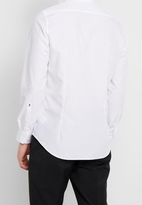 Witte langesleevenshirt met knoopmanchetten, gladde stof, getailleerde pasvorm en een afgeronde onderkant. Kenmerkt zich door subtiele stikseldetails op de mouwen.