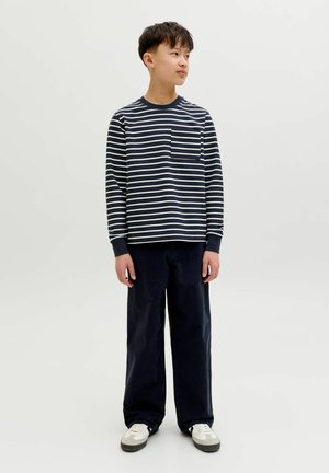 Dreng stående og kiggende mod højre, iført en navy og hvidstribet langærmet skjorte, navy bukser med vide ben og hvide sneakers.