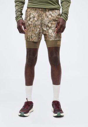 Mann iført olivengrønn langermet skjorte, blomstrete og kamuflasjemønstrete shorts, olivengrønne kompresjonsshorts, hvite sokker og burgunder løpesko.