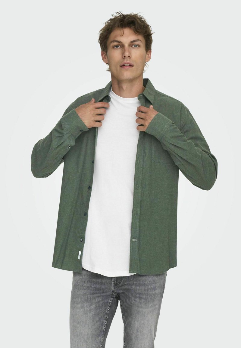 Chemise verte à manches longues, en tissu texturé, portée déboutonnée par-dessus un T-shirt blanc. Coupe slim et jeans gris visibles en dessous.