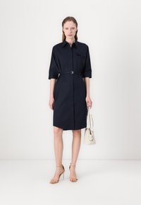 Navy shirt dress with a belted waist, short sleeves, collar, and a chest pocket. Smooth fabric, knee-length cut, paired with a light handbag.
