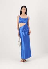 Vestido azul de cetim com um recorte na cintura, alças finas e uma saia longa com fenda. Modelo segurando uma pequena bolsa clara.
