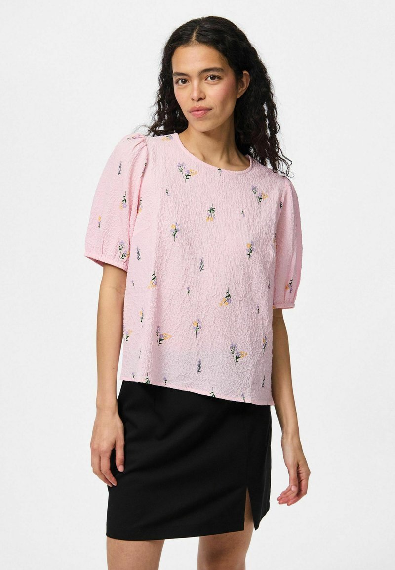 Top corto a maniche corte di colore rosa con una texture e ricami floreali, abbinato a una gonna nera con uno spacco laterale. Scollo rotondo semplice, vestibilità comoda.