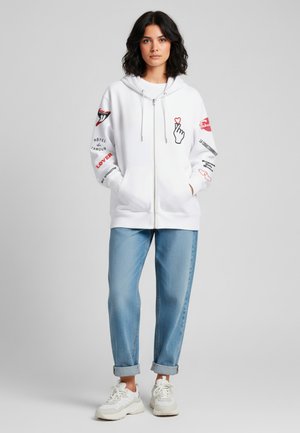 Felpa bianca con zip e stampe grafiche sulle maniche, che presentano cuori e testo. Abbinata a jeans azzurri arrotolati e sneakers bianche.
