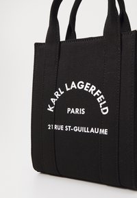 Zwarte canvas draagtas met korte handvatten, met witte tekst: "KARL LAGERFELD PARIS 21 RUE ST-GUILLAUME." Minimalistisch ontwerp, stevig materiaal.