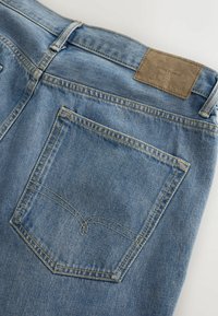 Zone de la poche arrière d'un jean denim bleu clair avec des coutures doublement piquées et un patch en cuir marron portant l'étiquette "DENIM SUPPLY".
