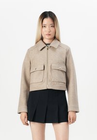 Cropped beige wool jacket with a collar, two front pockets, and a zip closure. Worn over a black pleated skirt. Smooth texture.