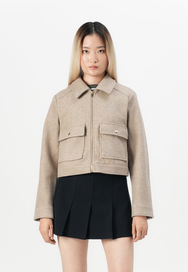 Cropped beige wool jacket with a collar, two front pockets, and a zip closure. Worn over a black pleated skirt. Smooth texture.