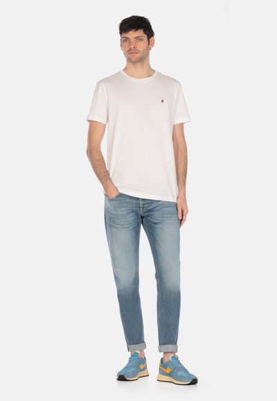 T-shirt bianca in cotone con maniche corte e un piccolo logo nero, abbinata a jeans in denim azzurro e sneakers blu con accenti arancioni.