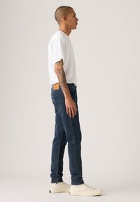 Balti medvilninė marškinėliai, mėlyni džinsai iš šono. Džinsuose gale yra odinė dekoratyvinė detalė, o batai – drobiniai sportbačiai su apvalia pirštine.