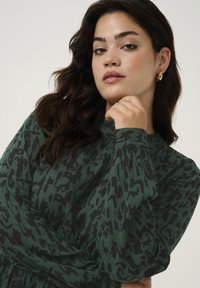 Robe vert foncé avec un motif abstrait noir, manches longues et col haut. La texture semble douce et légèrement côtelée. Boucles d'oreilles en or visibles.