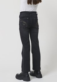 Persona vestida con jeans de mezclilla negros con costuras en espiral en los bolsillos traseros, botas negras gruesas y una camiseta blanca frente a un fondo liso.