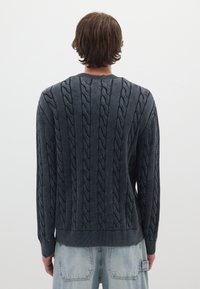 Dunkelgrauer Kabelstrickpullover mit einem gerippten Halsausschnitt und Bündchen, der vertikale Texturmuster aufweist. Kombiniert mit hellblauen Jeans.