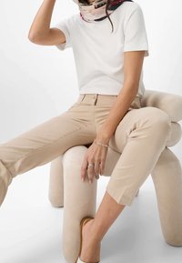 T-shirt blanc à manches courtes, pantalon beige ajusté, anneaux en argent simples, assis sur un tabouret beige doux et courbé, avec une écharpe à motifs autour du cou.