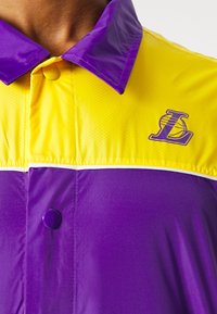 Primer plano de una chaqueta amarilla y morada con un botón a presión y un logo deportivo que presenta una "L" estilizada y un balón de baloncesto en el pecho.
