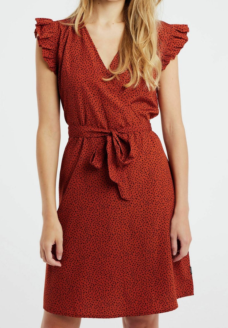 Femme portant une robe sans manches, de couleur rouille avec des taches noires, des épaules froncées, un décolleté en V et une ceinture nouée à la taille.