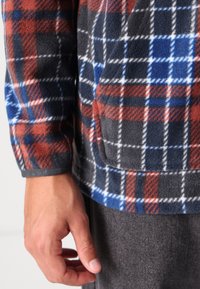 Fleece shirt in plaid pattern with red, blue, and gray hues. Features a front pocket and ribbed cuffs. Soft texture, warm and cozy design.