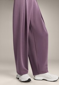 Lässige, weiche lila Hosen mit glatter Textur und plisseeartigem Design, kombiniert mit weißen Sportschuhen.