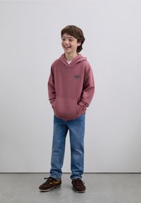 Bordowa bluza z kapturem z przednią kieszenią i logo, zestawiona z niebieskimi dżinsami i brązowymi trampkami. Tkanina wygląda na miękką, o casualowym kroju.