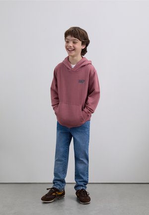 Felpa con cappuccio color marrone scuro con una tasca frontale e logo, abbinata a jeans blu e sneaker marroni. Il tessuto sembra morbido con una vestibilità casual.