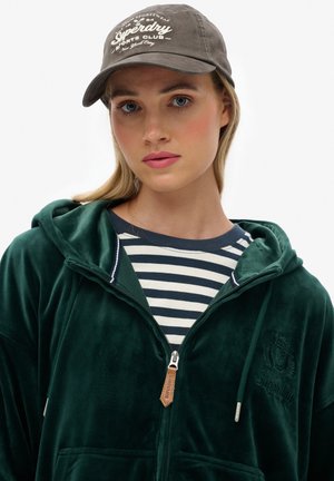 Green velvet zip-up hoodie with a logo, paired with a navy and white striped shirt. A brown cap with "Superdry Sports Club" text is worn.