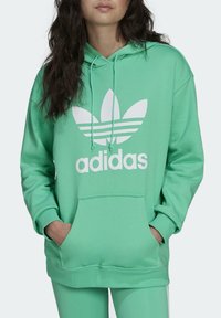 Mátově zelená mikina s kapucí s bílým logem Adidas a listovým motivem. Obsahuje přední kapsu a stahovací šňůrku na kapuci. Měkký materiál.