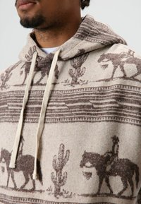 Beige capuchontrui met bruine zuidwestelijke patronen met cactussen en ruiters. Gevoerd materiaal met contrasterende trekkoorden.