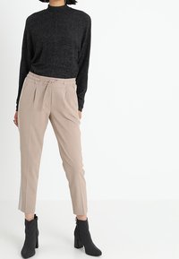 Svart långärmad topp med hög hals, beige avsmalnande byxor med dragsko i midjan och svarta randiga ankelskor.