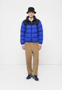 Blå og sort pufferjakke med lynlås foran, ribbede manchetter og sidelommer. Sammen med beige bukser og blå sneakers. Sort bøttehue bæres.
