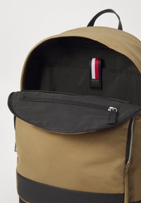 Zaino marrone con una tasca nera con zip, interno in tessuto a righe e un'etichetta in pelle. Materiale liscio e design strutturato.