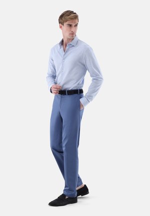 Junger Mann, der ein hellblaues Hemd mit Knöpfen, blaue Hose, schwarzen Gürtel und schwarze Loafers trägt und mit einer Hand in der Hosentasche steht.