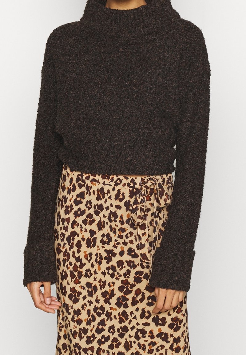 Personne portant un pull texturé marron foncé et une jupe beige avec un imprimé léopard marron foncé et orange, nouée sur le côté avec un nœud.