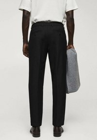 Homme portant un pantalon de costume noir, une chemise blanche à manches courtes, des chaussures foncées, tenant un pull en tricot gris plié dans la main droite, debout devant un fond uni.