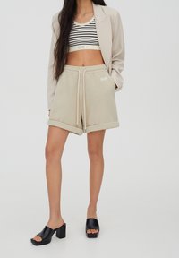 Shorts en polaire beige avec ourlet replié, taille à cordon de serrage et détail de logo blanc, associés à un crop top à rayures noires et blanches et des mules à talons noirs.