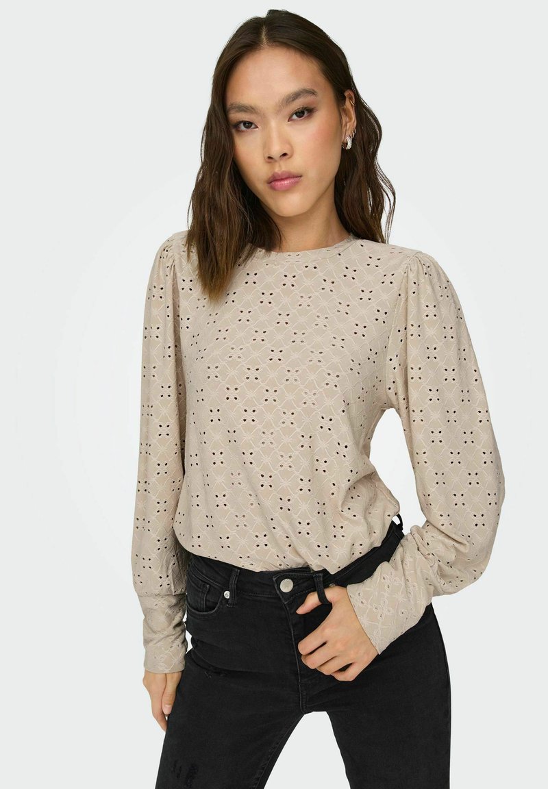 Beige langærmet bluse med hulmønstre, rund halsudskæring og puffede ærmer, parret med sorte jeans.