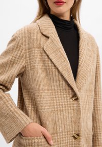 Cappotto di lana a scacchi beige con collo a revers, due tasche frontali e due bottoni. Tessuto morbido con un motivo a quadretti.