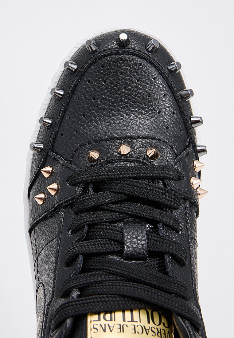 Zapatilla negra de cuero con superficie texturizada, que cuenta con tachuelas metálicas y cordones negros. Suela transparente con marca visible en la plantilla.