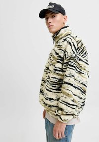 Fleece-Pullover mit hohem Kragen im cremefarbenen und schwarzen Tigermuster. Verfügt über eine flauschige Textur und weite Ärmel, kombiniert mit hellblauer Jeans.