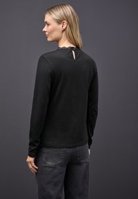 Frau mit blonden Haaren trägt ein schwarzes Langarmoberteil mit Spitzenkragen und Schlüsselloch-Rücken, kombiniert mit abgedunkelten schwarzen Jeans vor einem grauen Hintergrund.