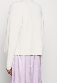 Weißer, gerippter Strickpullover mit lässiger Passform und überschnittenen Schultern. Kombiniert mit einem fliederfarbenen, glänzender texturierten Rock, der einen geschichteten Stil zeigt.