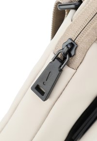 Zaino beige in materiale con finitura testurizzata, dotato di un tirante della cerniera nera con un dettaglio a forma di punto esclamativo e una robusta tracolla color terra.