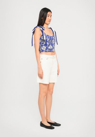 Woman in floral blue crop top with shoulder ties, white high-waist shorts, and black ballet flats standing sideways on plain background.