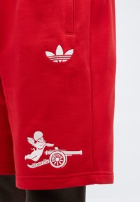 Červená textília s bielym vyšívaným logom Adidas nad anjelom strieľajúcim z dela, zobrazená na odeve nosenom na nohe.