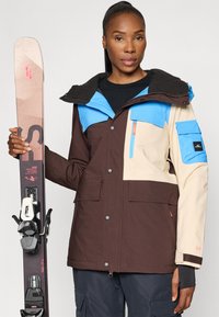 Ski jakna u boji smeđe, krem i plave, sa kapuljačom, više džepova i kopčama na dugmad. Model drži par skija.