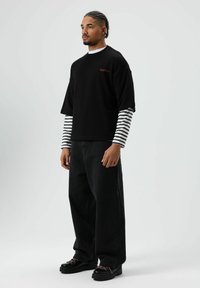 T-shirt noire surdimensionnée avec broderie rouge, portée par-dessus une chemise rayée à manches longues. Portée avec un jean noir à jambes larges et des chaussures épaisses noires.