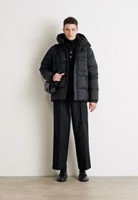 Zwarte pufferjack met een gestikte ontwerp, lange mouwen en een capuchon; zwarte coltrui; losse zwarte broek; zwarte schoenen; schoudertas.